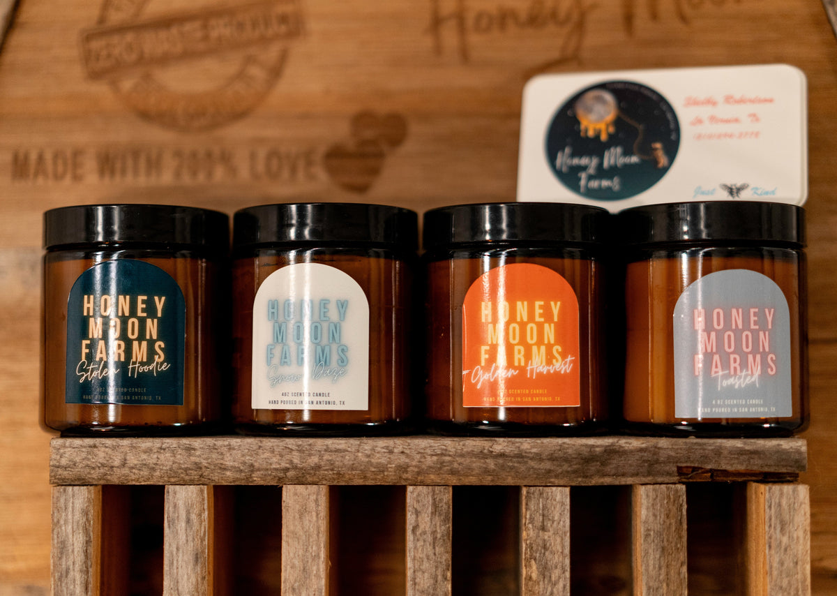 A set of four hand-poured beeswax candles in various colors, each labeled with different scent names such as 'Honey Farms' and 'Moon Pies'. The candles are displayed in a wooden container with a rustic label that says 'Made with 200% Love'.