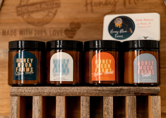 A set of four hand-poured beeswax candles in various colors, each labeled with different scent names such as 'Honey Farms' and 'Moon Pies'. The candles are displayed in a wooden container with a rustic label that says 'Made with 200% Love'.