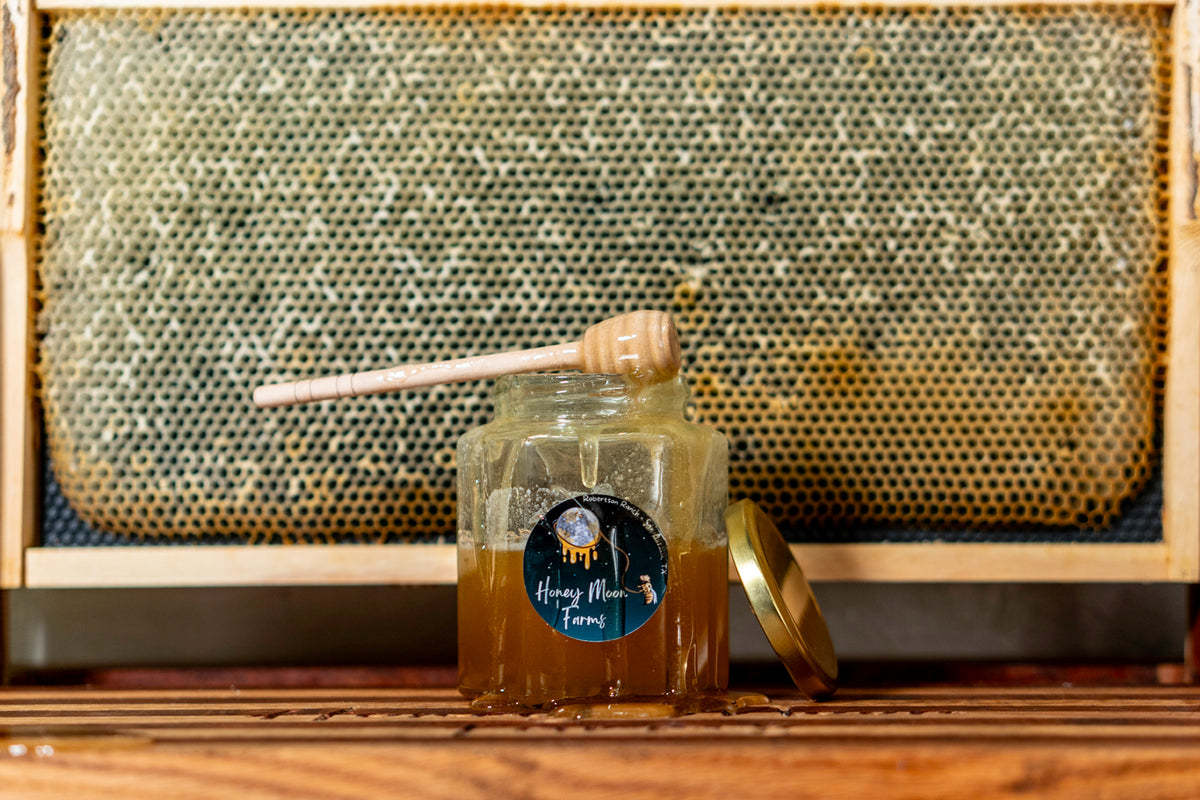 A jar of Honey Moon Farms Raw Texas honey placed in front of a honeycomb, with a label on the jar and a honey dipper.