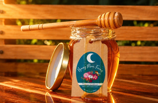 A jar of Mexican Wildflower honey with its lid off and a wooden honey dipper resting across the top, set on a wooden bench outdoors. The front label reads 'Honey Moon Farm' with a crescent moon and flowers.